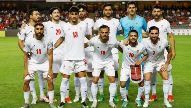 1775106104441_doi-tuyen-iran-1775101130375929895195 | MCG24 | موقع إلكتروني مغربي شامل متجدد على مدار الساعة Iran national football team players pose for a group photo on the field before a match, wearing white kits with red numbers; the goalkeeper is in a blue jersey.