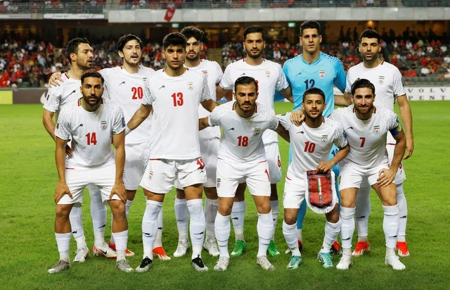 Iran national football team players pose for a group photo on the field before a match, wearing white kits with red numbers; the goalkeeper is in a blue jersey.