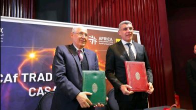 Two men in suits stand at a signing ceremony, each holding a colored folder (green and red) in front of a banner with Africa Development Club text on stage.