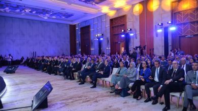 Large conference hall filled with formally dressed attendees seated in rows, watching a presentation on stage left, under blue and warm lighting.