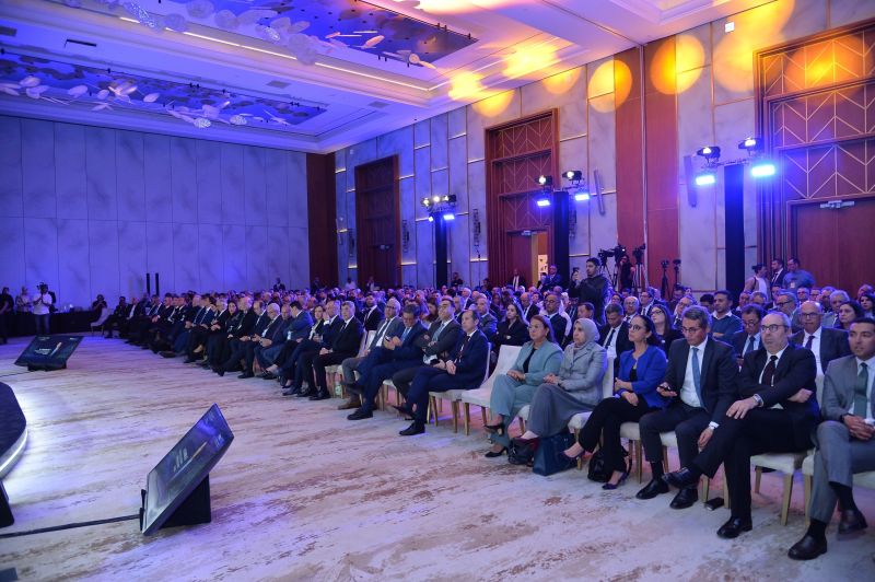 Large conference hall filled with formally dressed attendees seated in rows, watching a presentation on stage left, under blue and warm lighting.