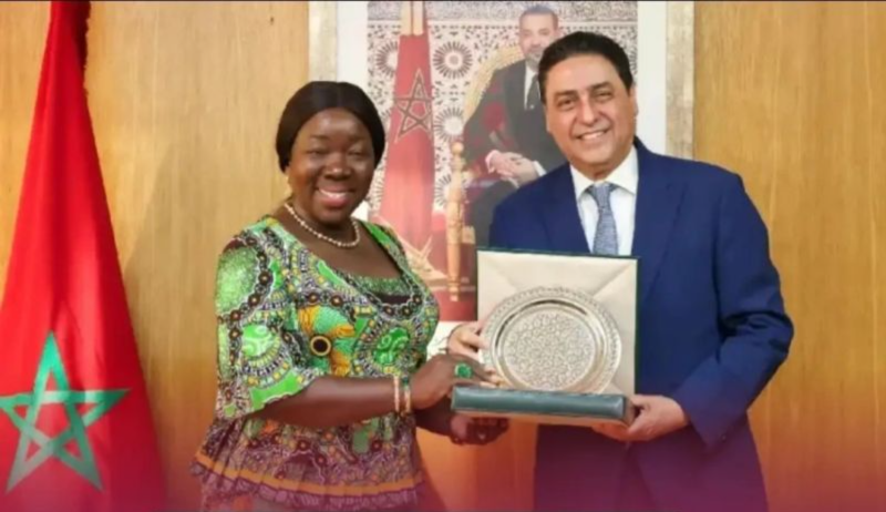 Two smiling people, a woman in a green patterned dress and a man in a blue suit, hold a ceremonial plaque together in front of a Moroccan flag and backdrop.