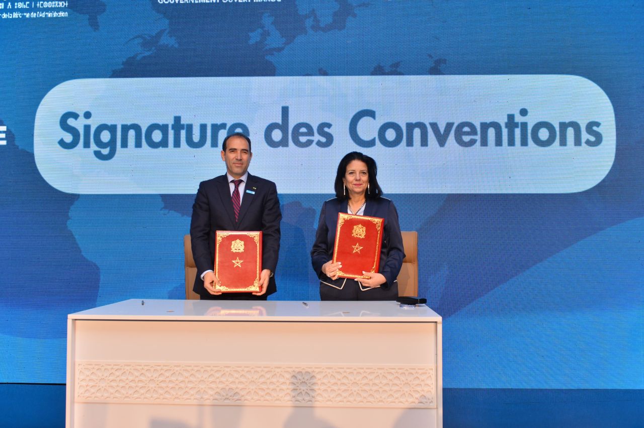 Two professionals in suits stand behind a desk, holding red ceremonial folders during a signing ceremony.