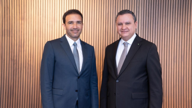 Two men in formal suits smiling, standing side by side against a vertical wooden slat wall.