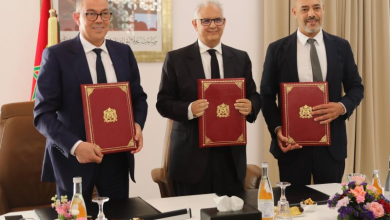 1778785871800 | MCG24 | موقع إلكتروني مغربي شامل متجدد على مدار الساعة Three men in dark suits stand at a conference table, each holding a red folder with a gold emblem during a formal ceremony.