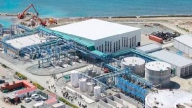 Aerial view of an industrial plant by the beach, featuring a large white-roofed building, blue piping, and storage tanks near the shoreline.