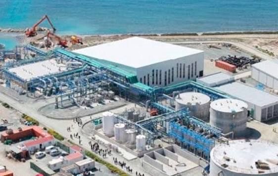 Aerial view of an industrial plant by the beach, featuring a large white-roofed building, blue piping, and storage tanks near the shoreline.
