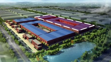 Aerial rendering of a large factory complex with blue and purple roofs, inner courtyards, and surrounding trees near a pond and roads.