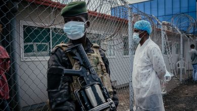 Masked soldier in camouflage and green beret standing near a barbed-wire fence outside a modular building, holding a rifle with equipment strapped to his chest.