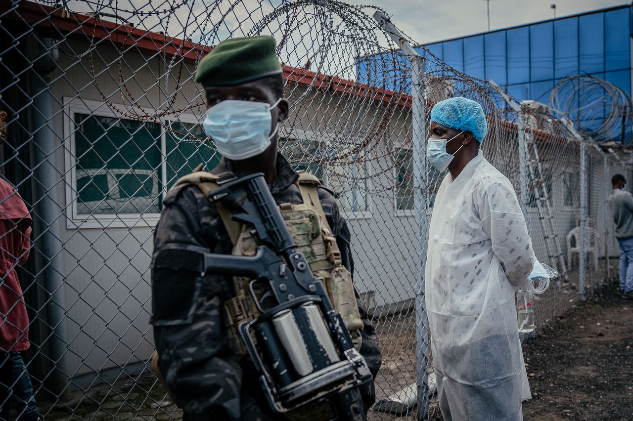 Masked soldier in camouflage and green beret standing near a barbed-wire fence outside a modular building, holding a rifle with equipment strapped to his chest.