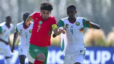 1779696223240hGpHNwebp-1779696223240_highres | MCG24 | موقع إلكتروني مغربي شامل متجدد على مدار الساعة Two soccer players sprint for the ball in a sunny match; one in a red jersey with number 7 and green shorts, wearing a yellow captain's armband.