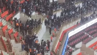 Large crowd of people in a sports arena, with some security stewards in orange vests guiding attendees up stairs near red seats.