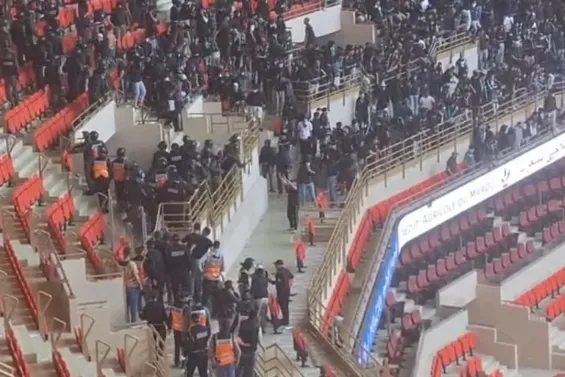 Large crowd of people in a sports arena, with some security stewards in orange vests guiding attendees up stairs near red seats.