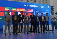 Group of men on a stage holding circular awards at an international event, with a blue backdrop displaying flags and the word NATIONALITES behind them.