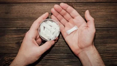 Hands hold a small circular container filled with white tablets on a wooden surface, with one tablet resting in the other open palm.