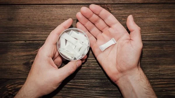 Hands hold a small circular container filled with white tablets on a wooden surface, with one tablet resting in the other open palm.