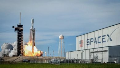 SpaceX rocket lifts off from a launch pad with a bright flame and plume, a tall gantry on the left and a large SpaceX hangar on the right.