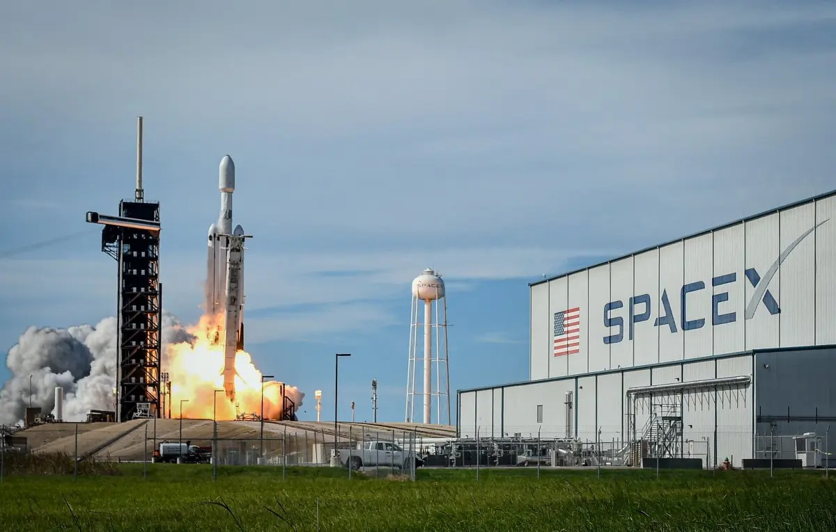 SpaceX rocket lifts off from a launch pad with a bright flame and plume, a tall gantry on the left and a large SpaceX hangar on the right.