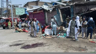 Group of civilians and men in traditional clothing picking through debris in a damaged market street after an explosion or attack, with a noticeable bloodstain on the pavement.