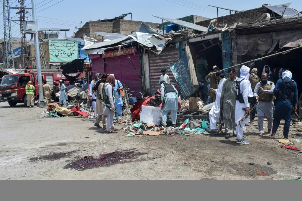 Group of civilians and men in traditional clothing picking through debris in a damaged market street after an explosion or attack, with a noticeable bloodstain on the pavement.