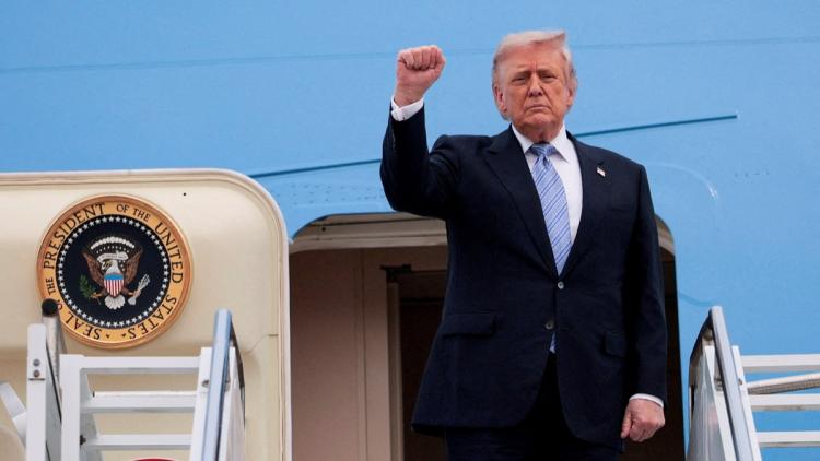 Former U.S. President Donald Trump raises his right fist while stepping down from Air Force One, with the presidential seal visible on the aircraft door.