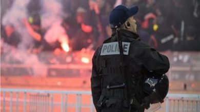 20260528114037125 | MCG24 | موقع إلكتروني مغربي شامل متجدد على مدار الساعة Police officer in tactical gear facing away from the camera, overlooking a crowd with flares and smoke in the background; vest reads POLICE.
