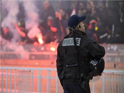 Police officer in tactical gear facing away from the camera, overlooking a crowd with flares and smoke in the background; vest reads POLICE.