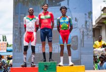 Three male cyclists stand on a winners' podium (gold, red, and green podiums) after a race, wearing their team jerseys as spectators look on.