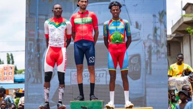 Three male cyclists stand on a winners' podium (gold, red, and green podiums) after a race, wearing their team jerseys as spectators look on.