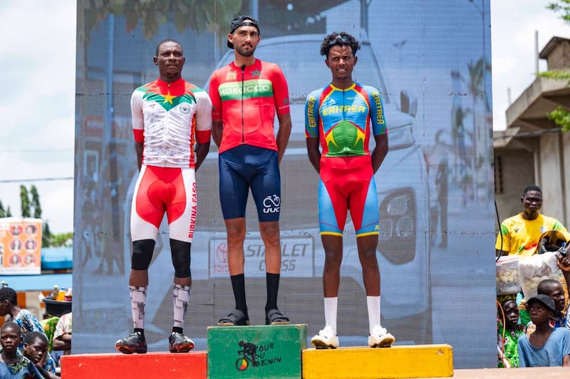 Three male cyclists stand on a winners' podium (gold, red, and green podiums) after a race, wearing their team jerseys as spectators look on.