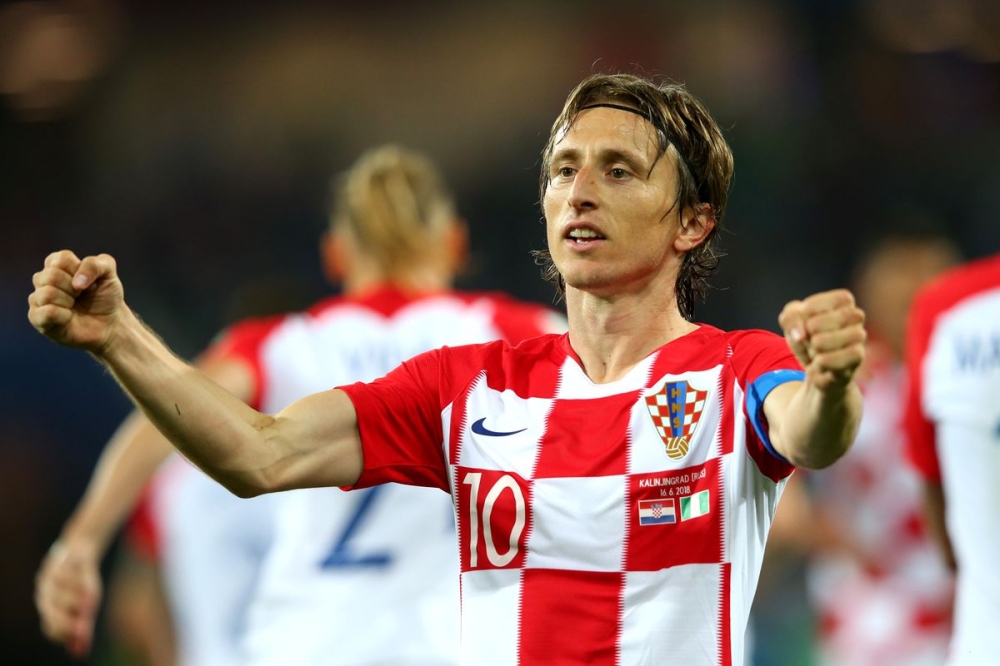 Croatian footballer in a red-and-white checkered jersey celebrates with arms raised on the field, wearing number 10 cap and captain armband.",
