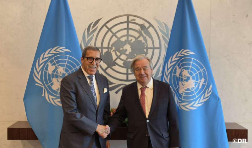 Two men in suits shake hands in front of United Nations flags and emblem, in a formal meeting setting.