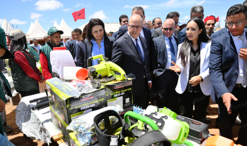 Group of officials and attendees at an outdoor expo inspecting bright green power tools on a display table under a blue sky.