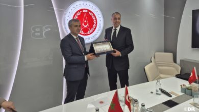 Two men in suits exchange a framed plaque in a conference room, with Moroccan flags on the table.