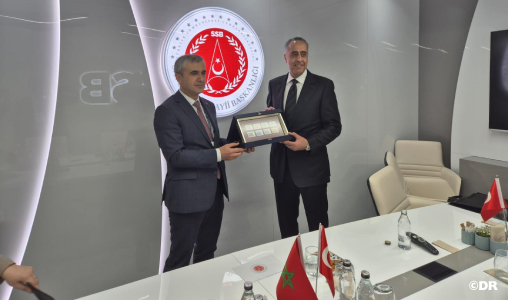 Two men in suits exchange a framed plaque in a conference room, with Moroccan flags on the table.