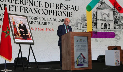 Man speaking at a podium during a conference on the network and Francophonie; Moroccan flag on the left and a framed portrait on an easel nearby.