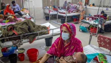 A woman in a pink sari wearing a mask cradles a baby while sitting beside a hospital bed in a crowded ward of patients and other families.