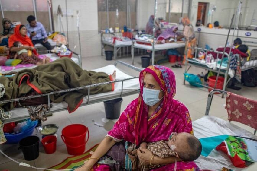 A woman in a pink sari wearing a mask cradles a baby while sitting beside a hospital bed in a crowded ward of patients and other families.