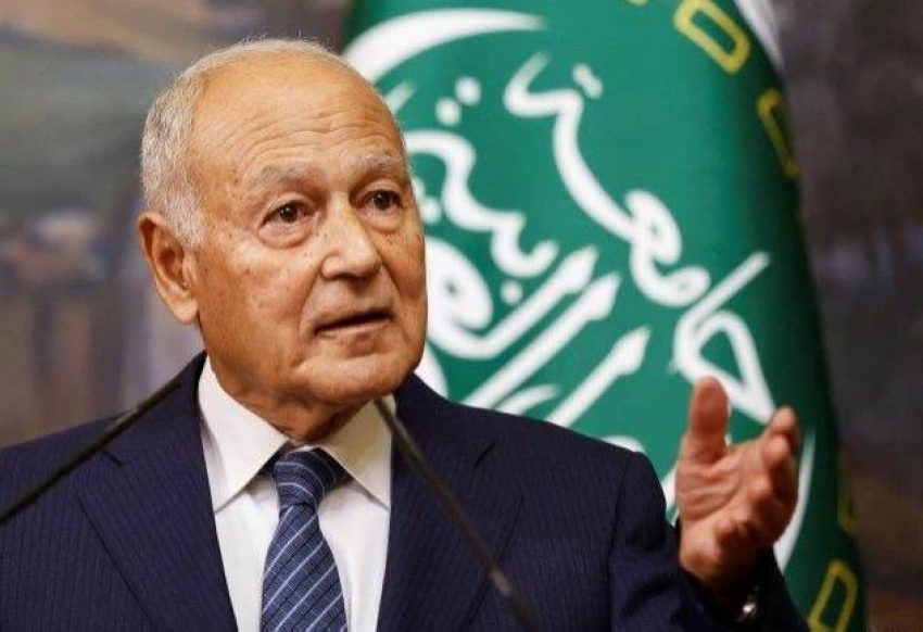 Older man in a dark suit and striped tie speaks at a podium with a green emblem behind him, addressing an audience.