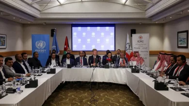 A formal meeting around a long U-shaped table with delegates, microphones, and water glasses, in a conference room with banners in the background and a projector screen showing a blue UN-related graphic.