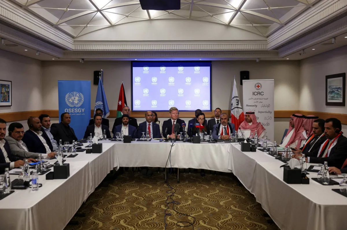 A formal meeting around a long U-shaped table with delegates, microphones, and water glasses, in a conference room with banners in the background and a projector screen showing a blue UN-related graphic.