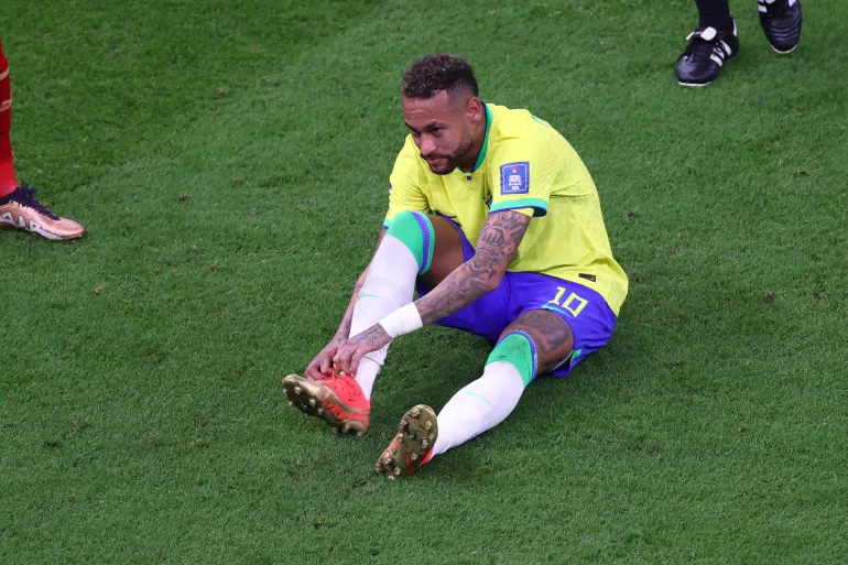 29538949 | MCG24 | موقع إلكتروني مغربي شامل متجدد على مدار الساعة Brazilian soccer player in a yellow kit sits on the grass and ties his red cleats during a match on a green field.