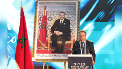 Man in a suit speaks at a podium, flanked by a framed portrait of the Moroccan king and the Moroccan flag behind him, at a formal event.