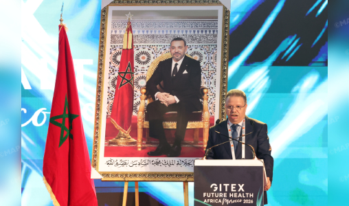 Man in a suit speaks at a podium, flanked by a framed portrait of the Moroccan king and the Moroccan flag behind him, at a formal event.