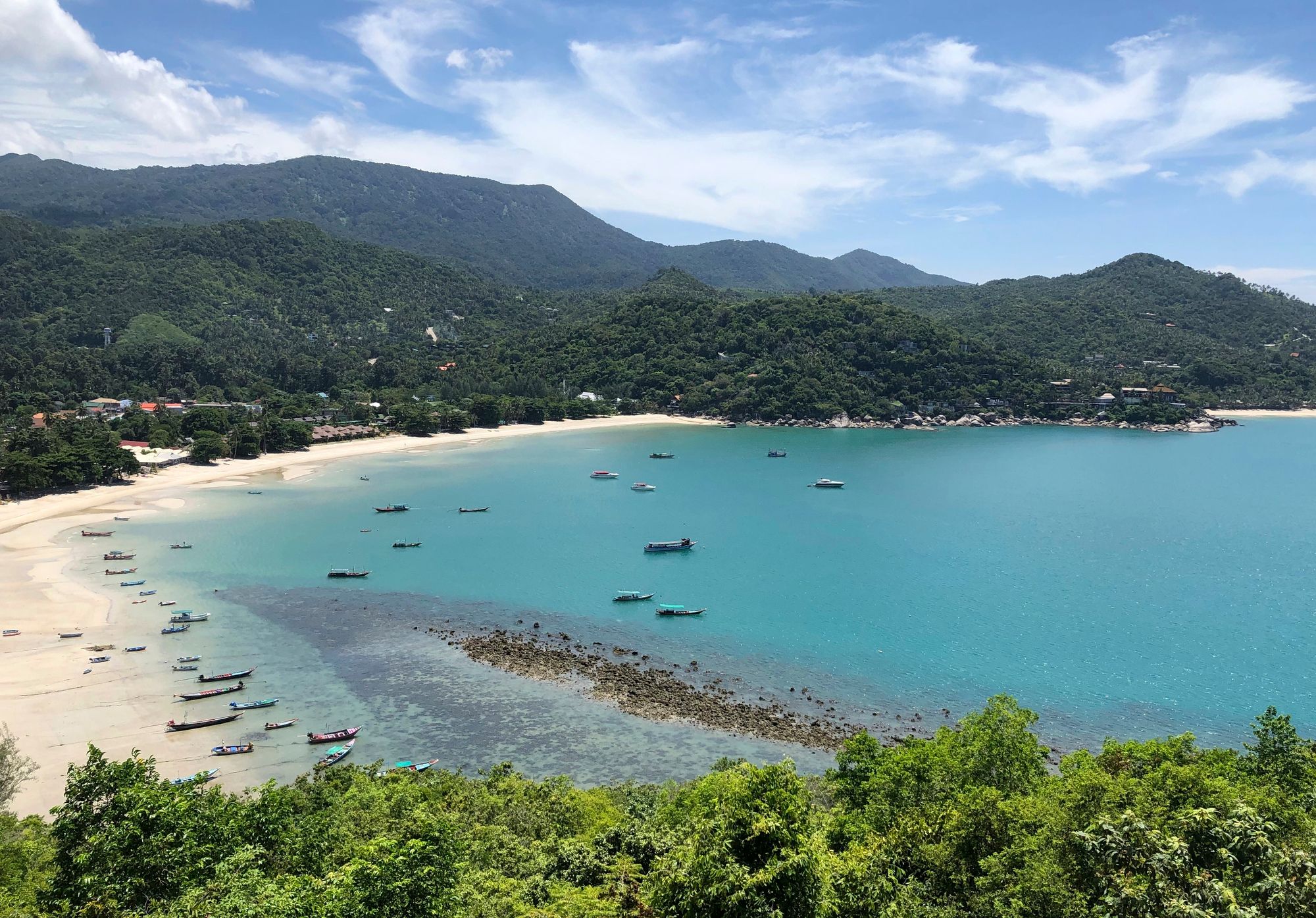 Coastal bay with a white-sand beach, turquoise water, boats anchored near shore, and forested hills in the background