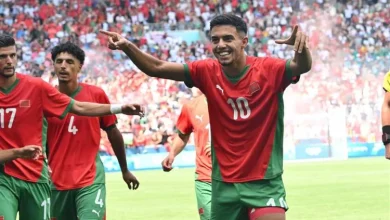 Soccer players in red and green uniforms celebrate on a sunny stadium field, with number 10 at the center raising his arms as teammates applaud and a cheering crowd behind them.