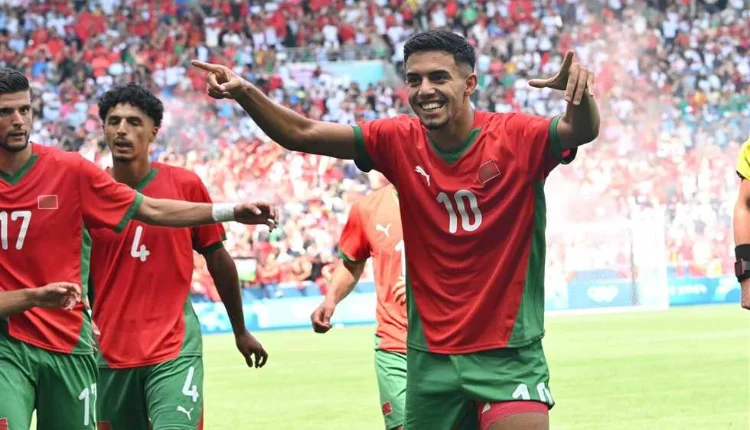 Soccer players in red and green uniforms celebrate on a sunny stadium field, with number 10 at the center raising his arms as teammates applaud and a cheering crowd behind them.