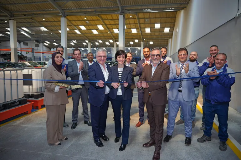 Group of professionals cutting a blue ribbon at a factory opening, with colleagues applauding in the background.