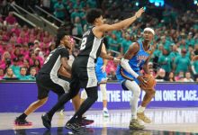 A basketball player in a teal Oklahoma City Thunder uniform dribbles the ball toward the basket while two defenders in black jerseys close in from the front, with a crowded arena in the background.