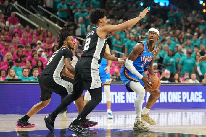 A basketball player in a teal Oklahoma City Thunder uniform dribbles the ball toward the basket while two defenders in black jerseys close in from the front, with a crowded arena in the background.